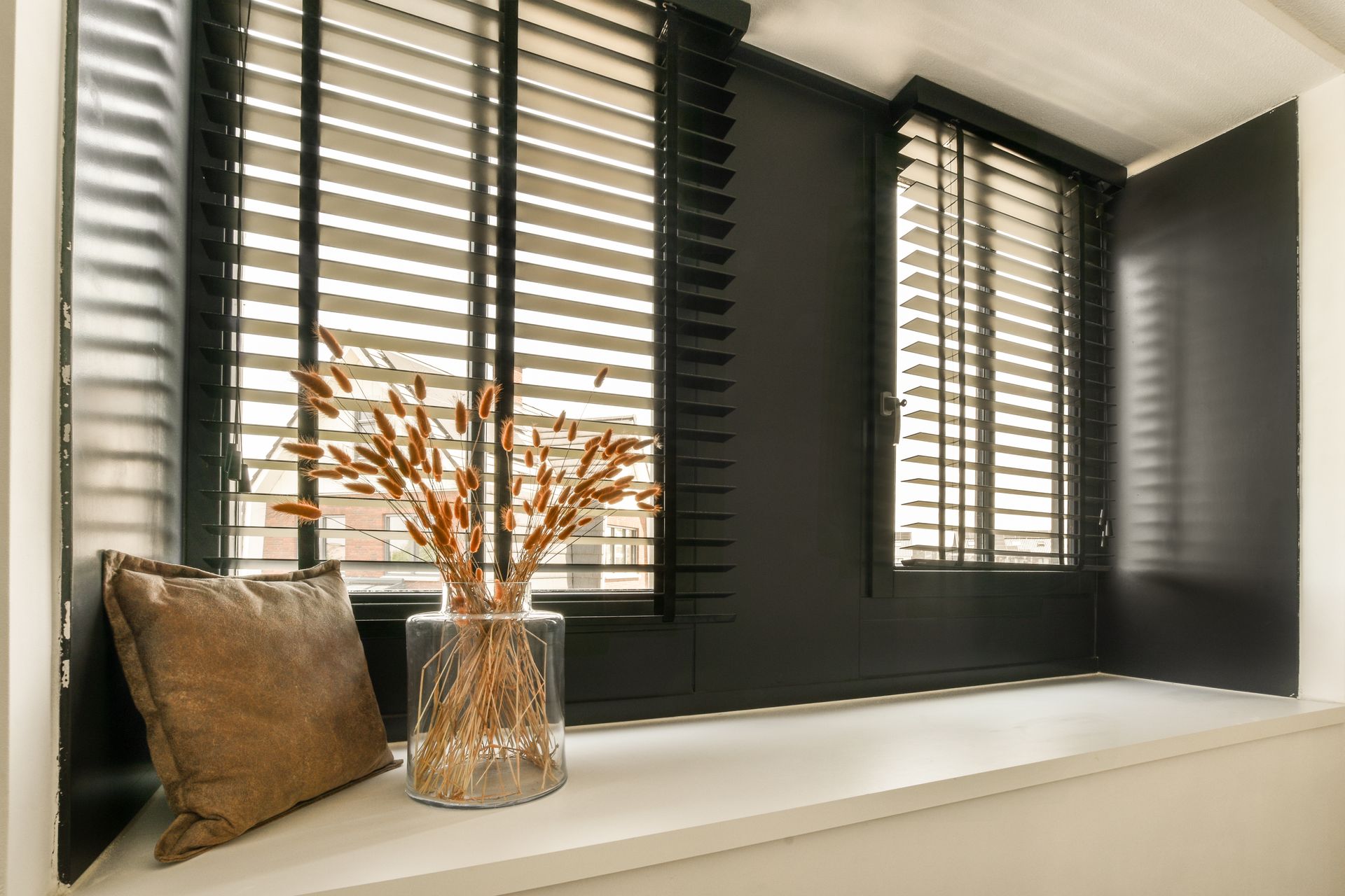 Un rebord de fenêtre avec un coussin marron et un vase en verre rempli de tiges de blé séchées, devant des stores noirs.