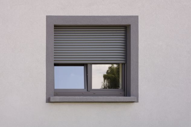 Une fenêtre carrée sur un mur blanc, avec un cadre gris, un volet partiellement fermé et une vitre réfléchissante.