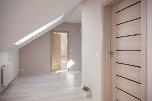 Un couloir lumineux avec un parquet clair, des murs beiges, un plafond mansardé avec un puits de lumière et deux portes en bois fermées.