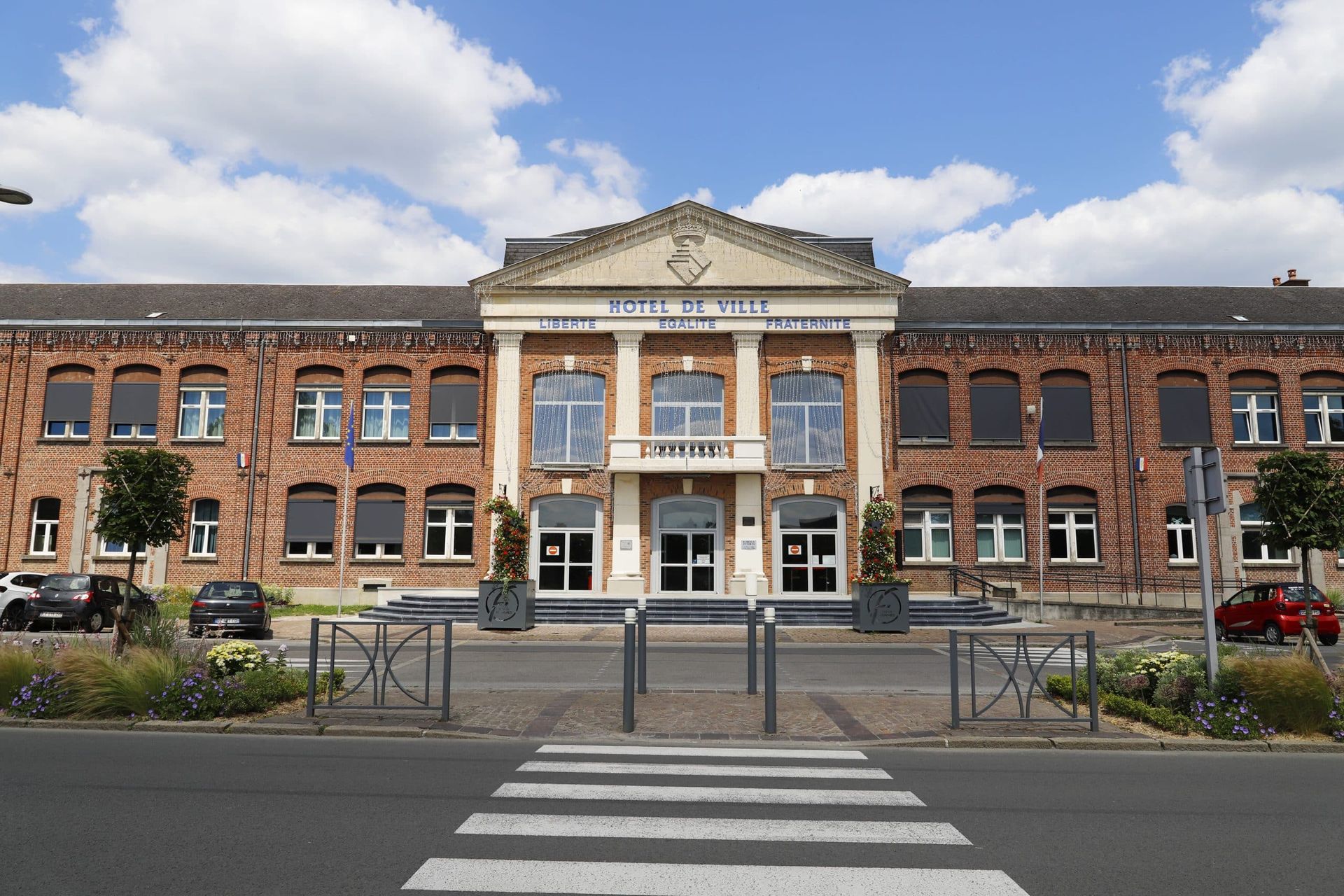 Vue d'ensemble à hauteur des yeux d'un hôtel de ville en briques de deux étages avec une entrée néoclassique, sous un ciel partiellement nuageux.