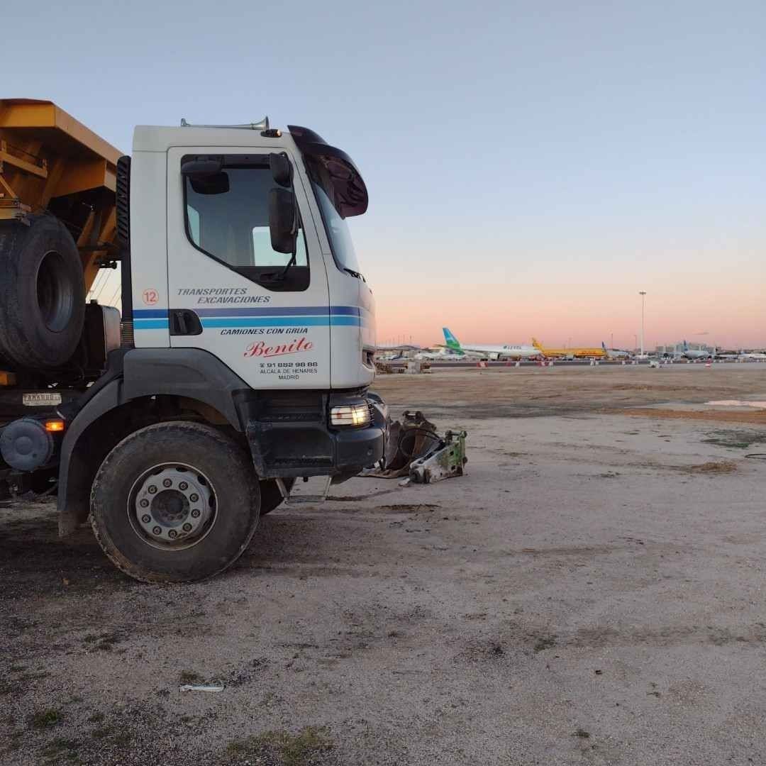Un camión volquete está estacionado en un campo de tierra con aviones al fondo.