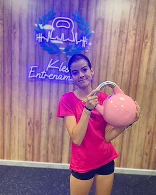 Una mujer con una camisa rosa sostiene una pesa rusa rosa en un gimnasio.