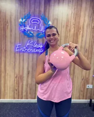 Una mujer con una camiseta sin mangas rosa sostiene una pesa rusa rosa en un gimnasio.
