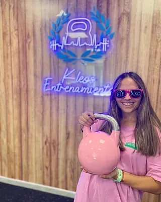 Una mujer sostiene una pesa rusa rosa frente a una pared de madera.