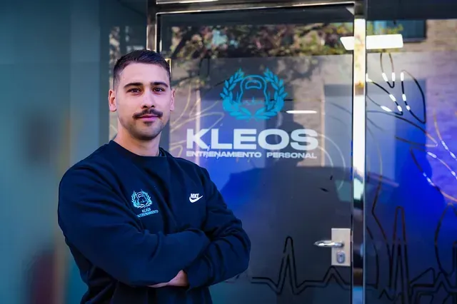 Un hombre con los brazos cruzados está parado frente a un cartel de Kleos.