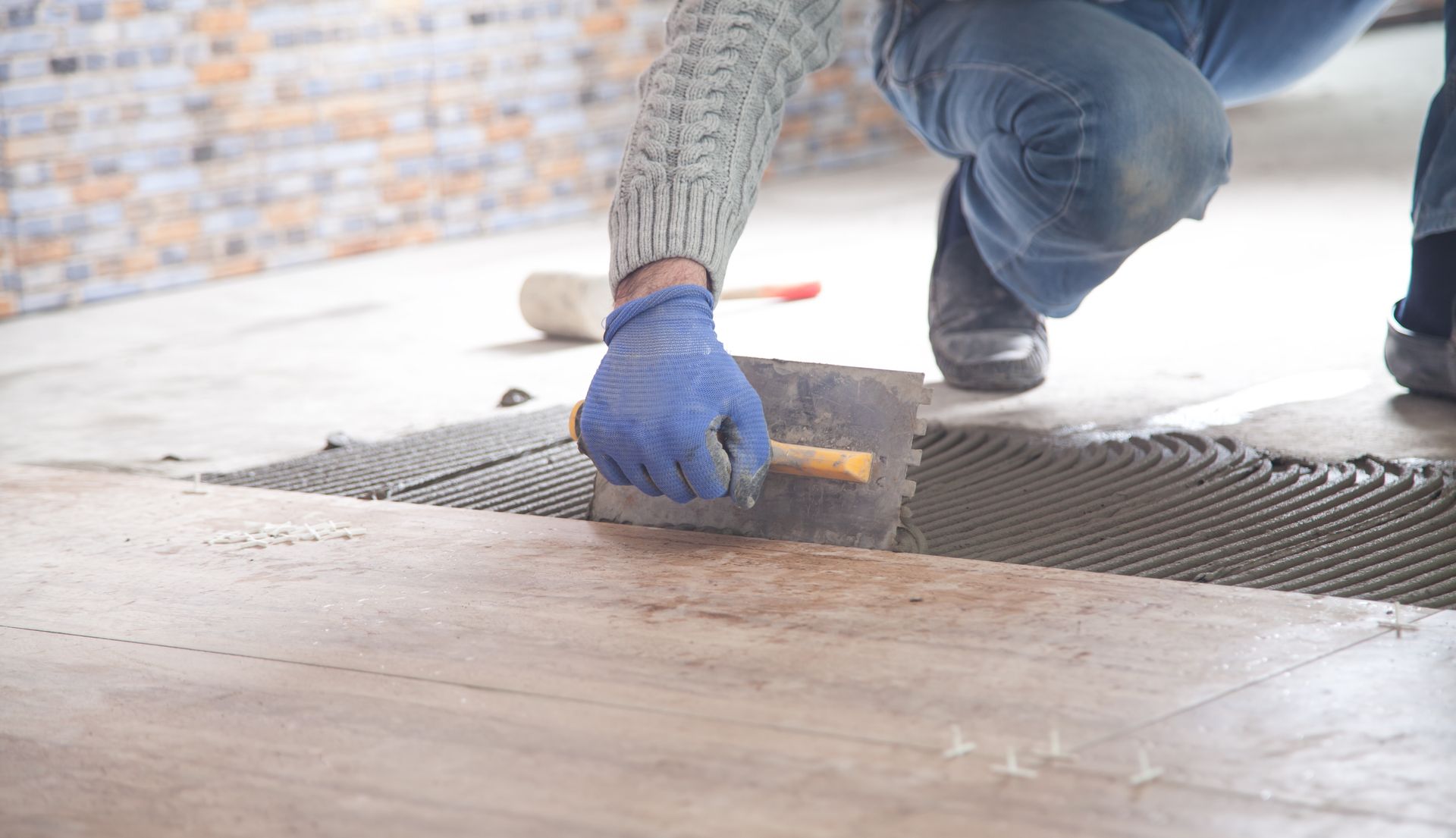 Un ouvrier étale du mortier sur un sol lors de la pose de carrelage, portant des gants bleus et un jean.