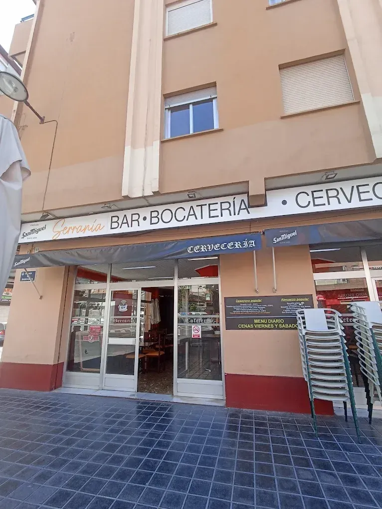 Fachada de un bar-cafetería con puertas de cristal, toldo a rayas y sillas apiladas sobre una acera embaldosada.