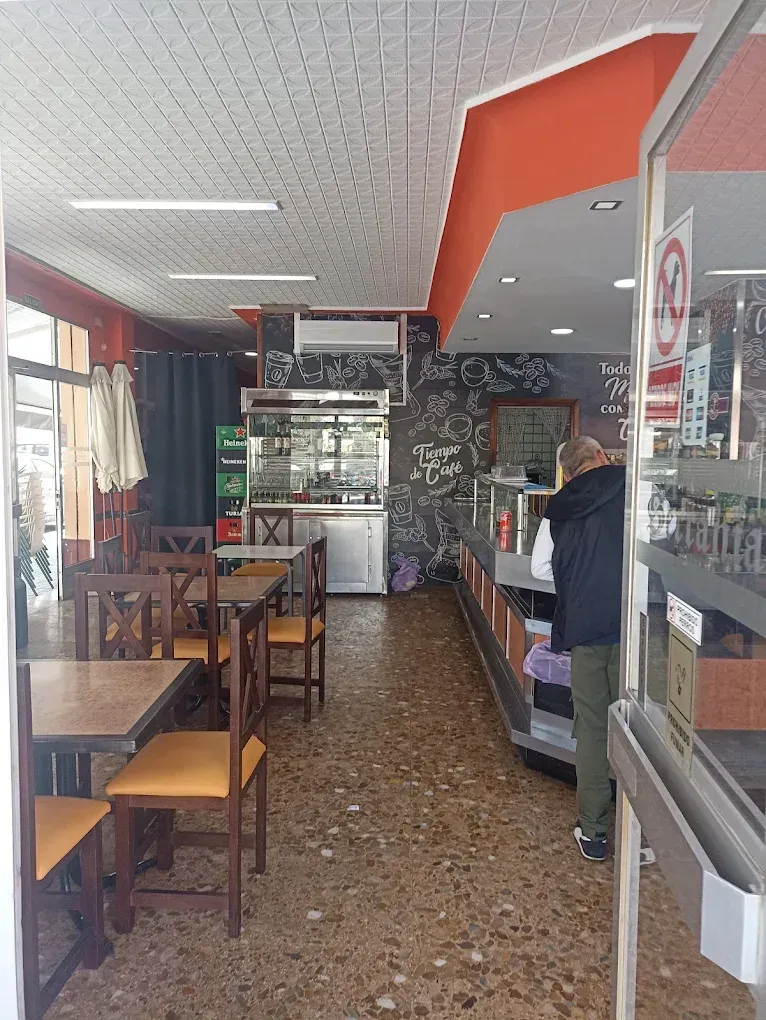 Interior estrecho de una cafetería con mesas, estantes con aperitivos y una persona detrás del mostrador.