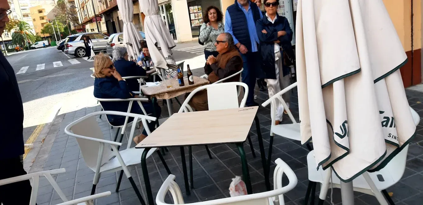 Mesas y sillas de café al aire libre en una acera soleada, con varias personas sentadas y de pie cerca.