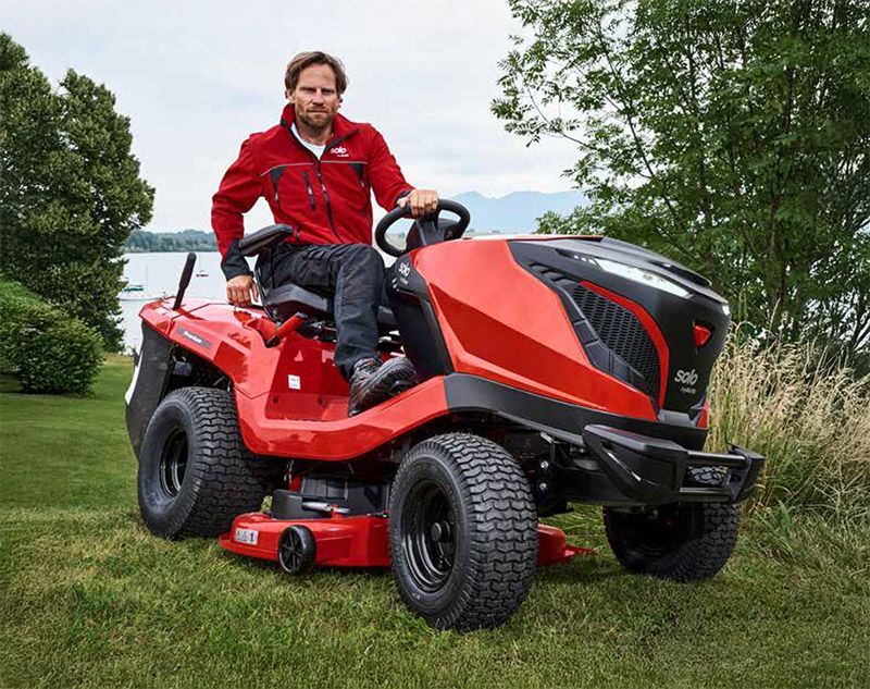 Mann in roter Jacke auf einem roten Rasentraktor, im Freien an einem See.