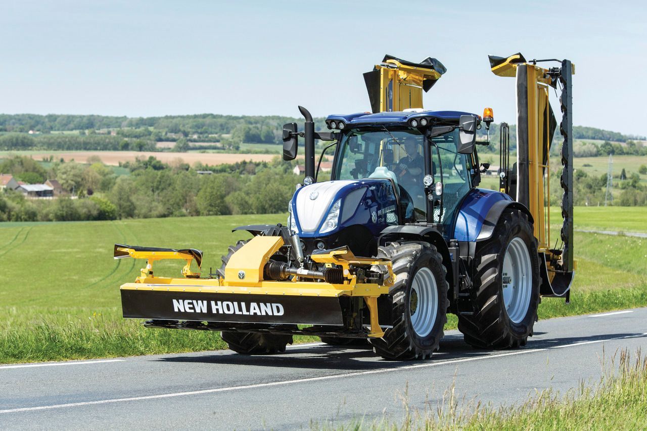 Blauer und gelber New Holland Traktor mit Mähwerk auf einer Straße in ländlicher Umgebung.