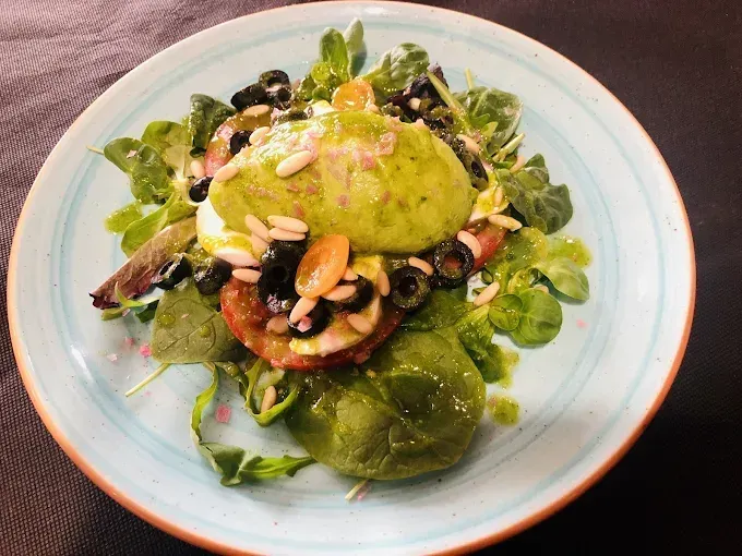 Ensalada con aguacate, tomate, aceitunas, piñones y aderezo, en plato azul.