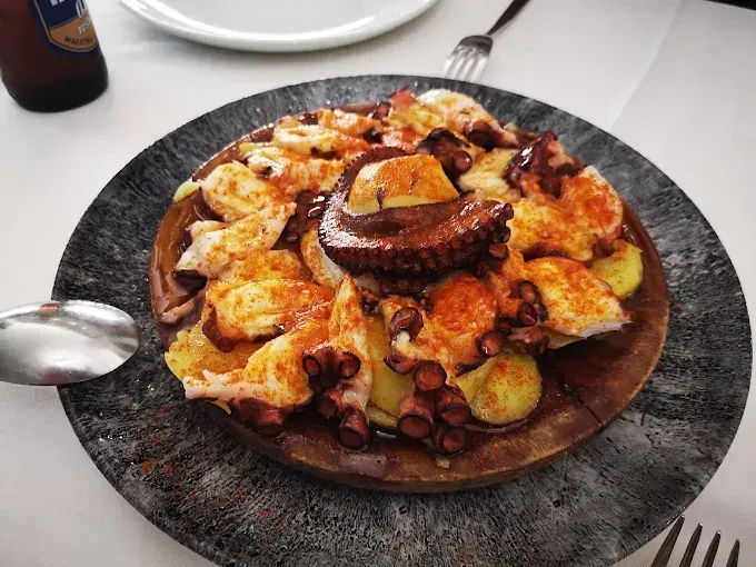 Plato de pulpo en plato de madera, cubierto con pimentón y patatas, servido sobre una superficie gris.
