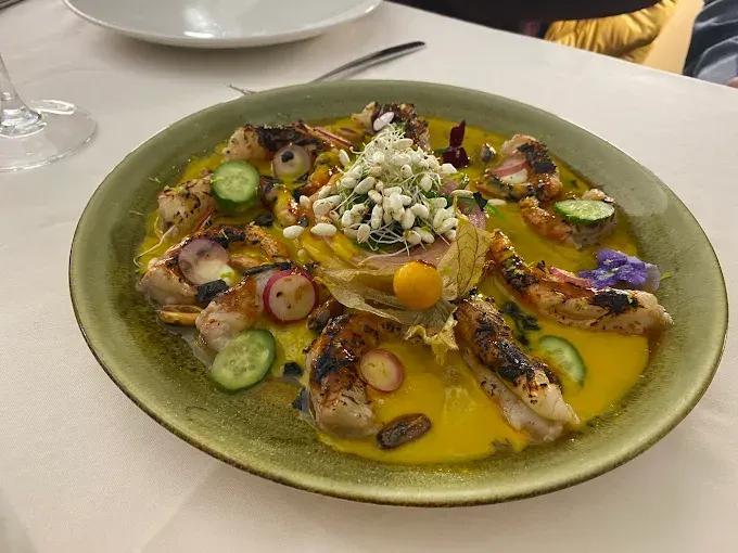 Plato de mariscos a la parrilla en salsa amarilla, adornado con verduras y una torre central de color blanco.