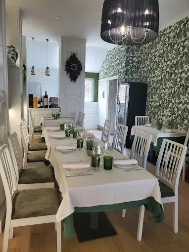 Comedor de restaurante con mesas largas, sillas blancas, detalles en verde y una pared con papel tapiz floral.