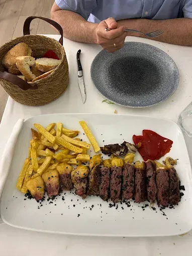 Cena de bistec con patatas fritas y pimiento rojo asado en un plato blanco. Cesta de pan y una persona sentada a la mesa.