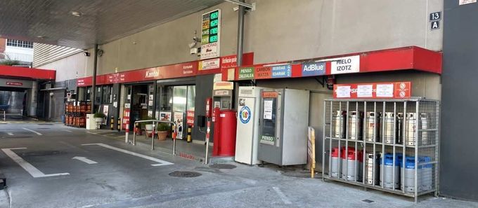 Exterior de una gasolinera con entrada de tienda, máquinas expendedoras y un estante de metal.