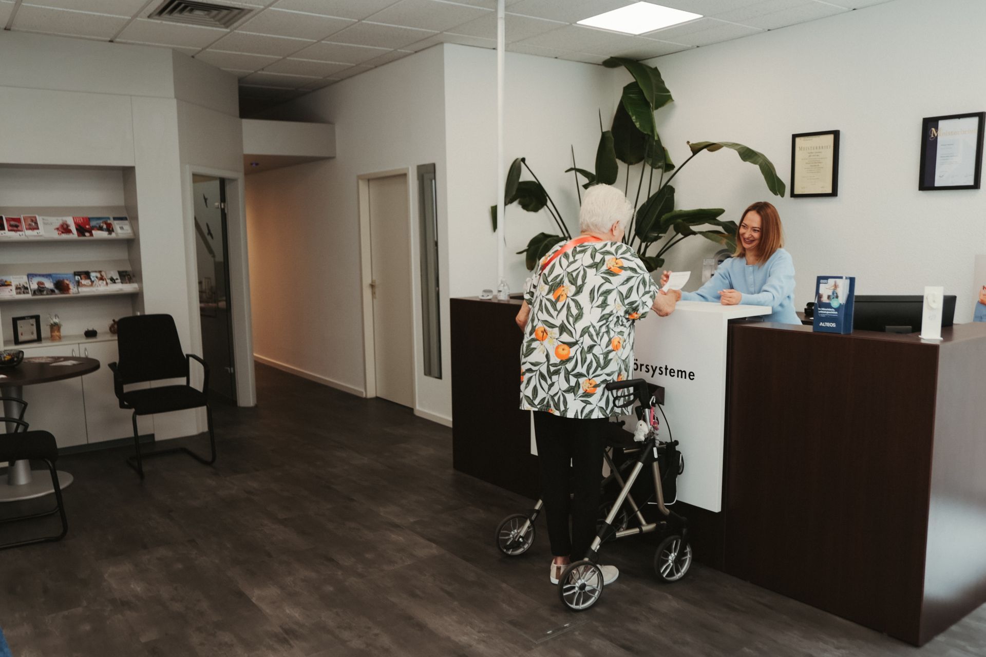 Empfangstresen in einer Bürolobby, an dem eine Person mit Rollator mit einer Rezeptionistin spricht.