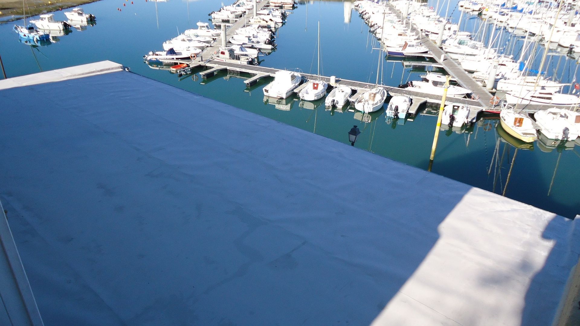 Vue aérienne d'un port de plaisance avec des bateaux blancs amarrés, de l'eau et un auvent bleu.