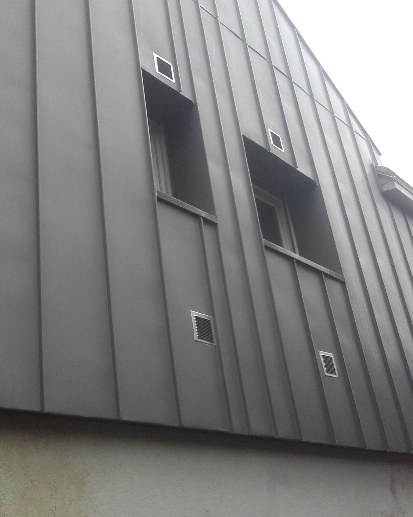 Bardage en planches beiges sur une maison au toit noir et aux accents de pierre ; on aperçoit également des gouttières et une petite fenêtre.