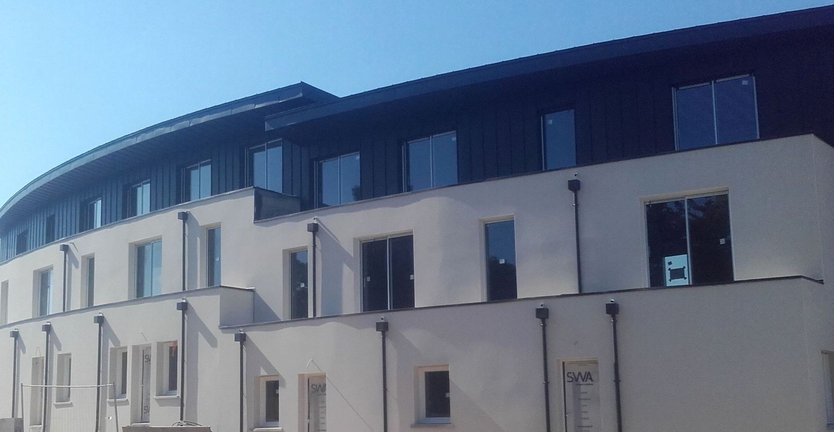 Bâtiment blanc de deux étages, au toit sombre et aux nombreuses fenêtres, sous un ciel bleu.