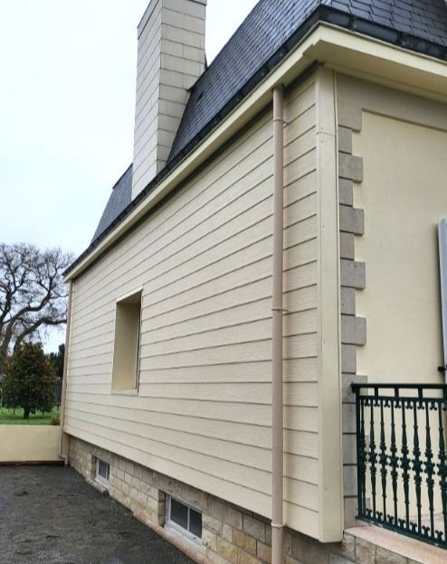 Un bâtiment avec un bardage beige, un toit noir, des boiseries beiges et une cheminée ; une partie d'une rambarde en fer forgé est visible.