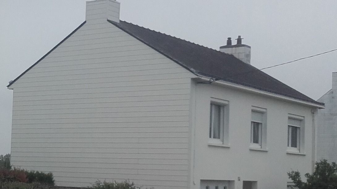 Maison blanche à deux étages avec toit noir, trois fenêtres, une cheminée et un ciel nuageux.
