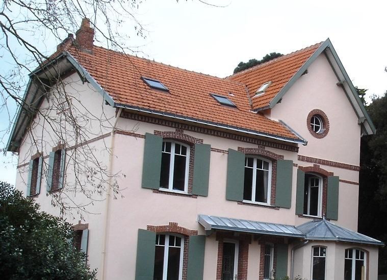 Maison rose avec toit de tuiles orange, volets verts, éléments en brique et un petit porche.