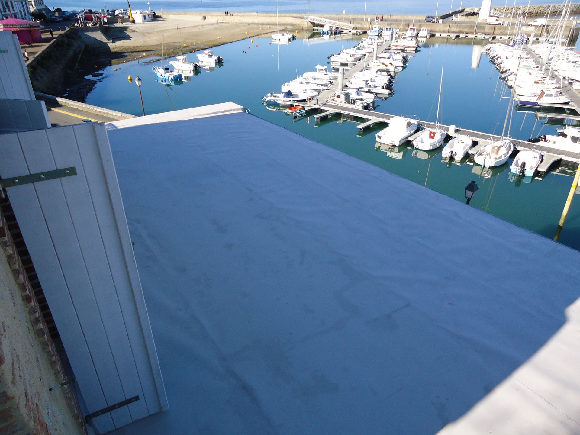 Vue aérienne d'un port de plaisance avec des bateaux blancs amarrés, de l'eau et un auvent bleu.