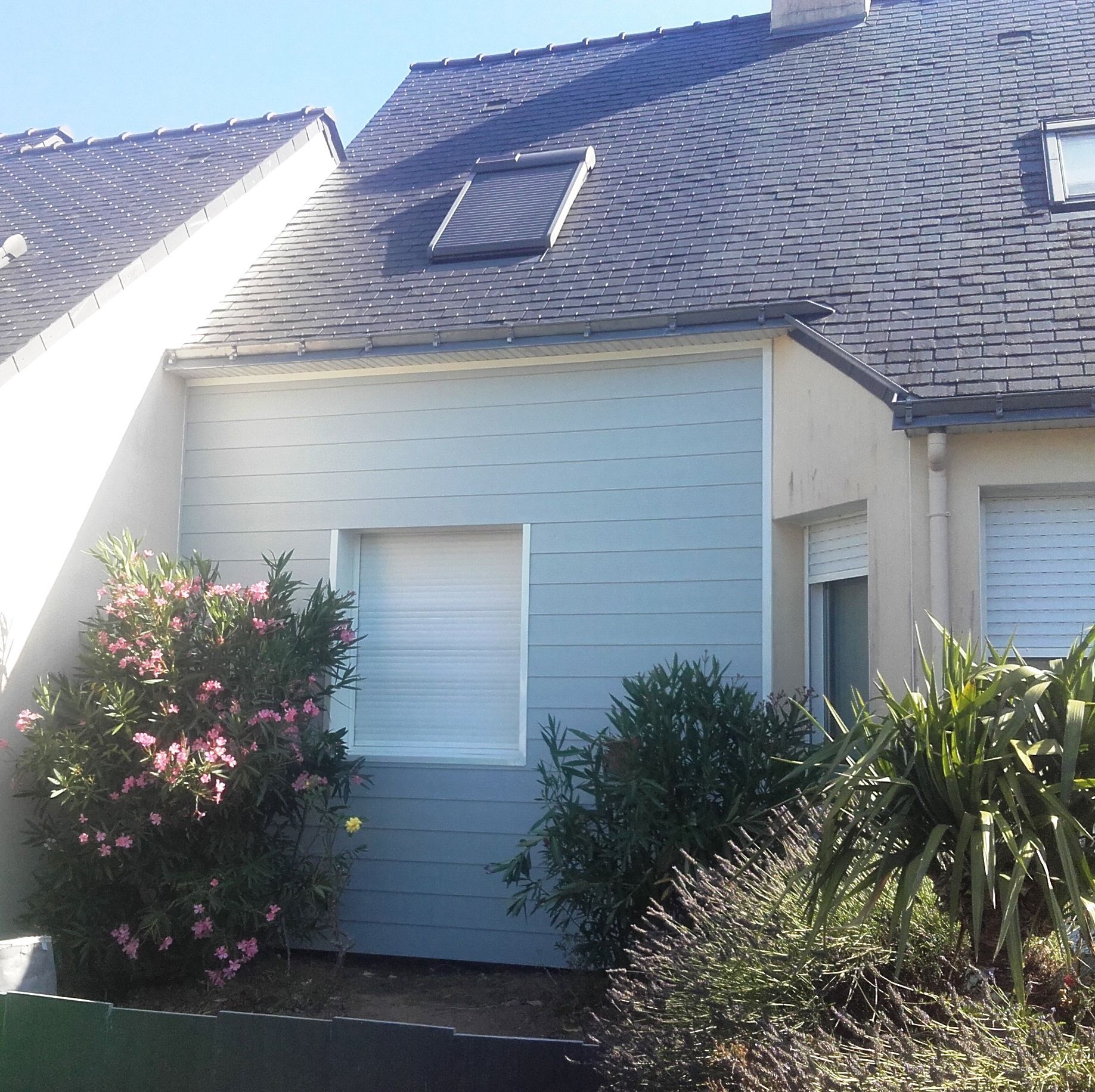 Bâtiment revêtu de métal gris, avec deux fenêtres et quatre aérations carrées. Des branches encadrent le côté.