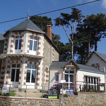 Maison au toit d'ardoise bleue, avec lucarnes et cheminée, se détachant sur un ciel bleu.