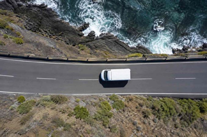 Una furgoneta blanca circulando por una carretera costera junto a las olas rocosas del océano, vista aérea.