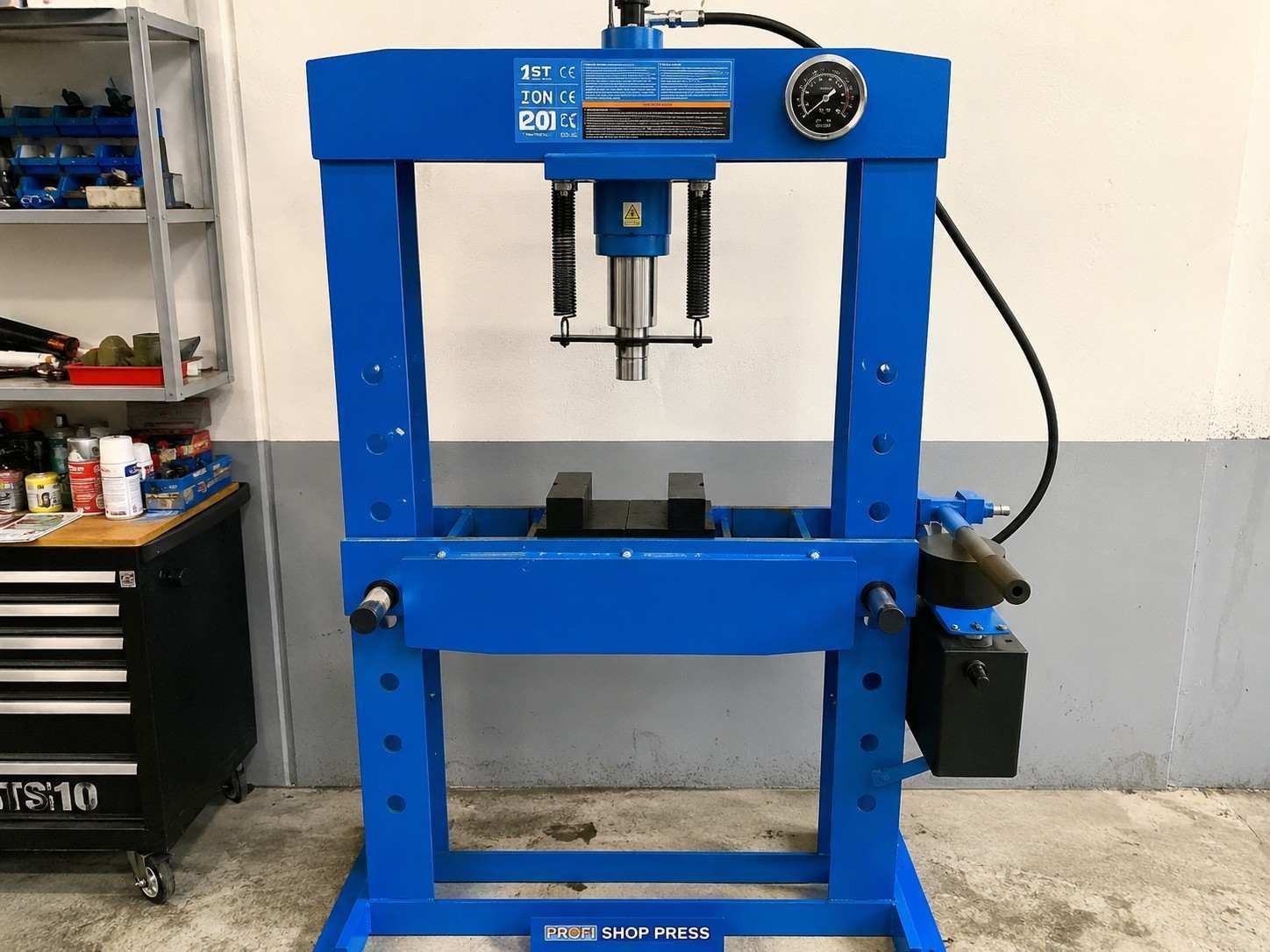 Prensa hidráulica azul en un taller mecánico, con manómetros y una plataforma de trabajo negra.