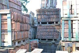 Plusieurs palettes en bois empilées avec des rangées de tuiles en terre cuite sont entreposées dans une zone extérieure.
