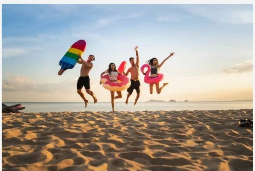 People jumping joyfully on a beach, holding inflatable floats and a popsicle, sunset in the background.