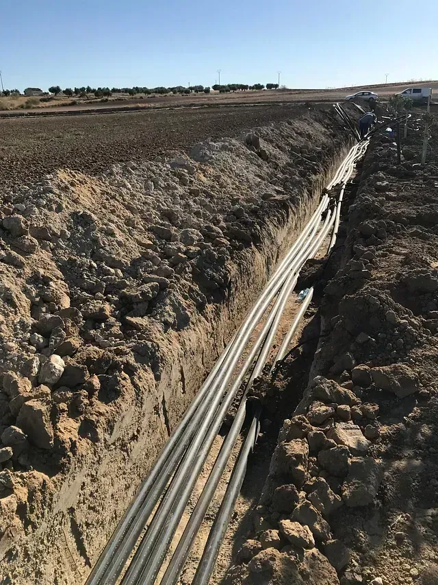 Zanja con múltiples cables grises tendidos en su interior, probablemente para servicios públicos subterráneos, en un campo rural y abierto.