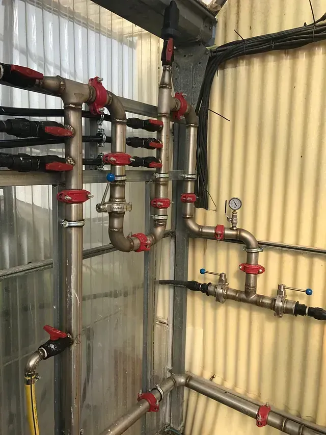 Una compleja red de tuberías metálicas con válvulas rojas y negras recorre una pared de metal corrugado. Algunas tuberías tienen mangueras amarillas conectadas.