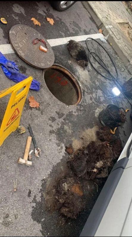 Alcantarilla abierta, señal de peligro, herramientas y escombros sobre el asfalto.