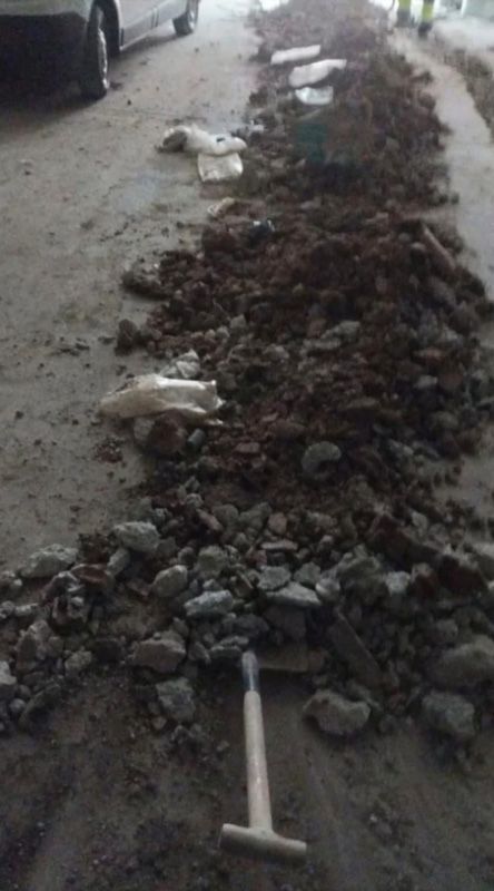 Zanja excavada en hormigón, llena de tierra y escombros; una pequeña pala se encuentra cerca, un vehículo al fondo.