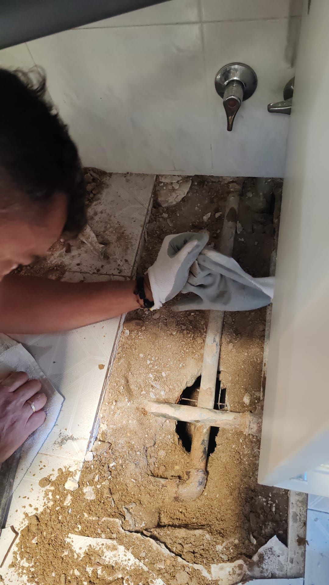 Una persona en un baño repara tuberías expuestas bajo baldosas dañadas. Guantes blancos, suelo beige.