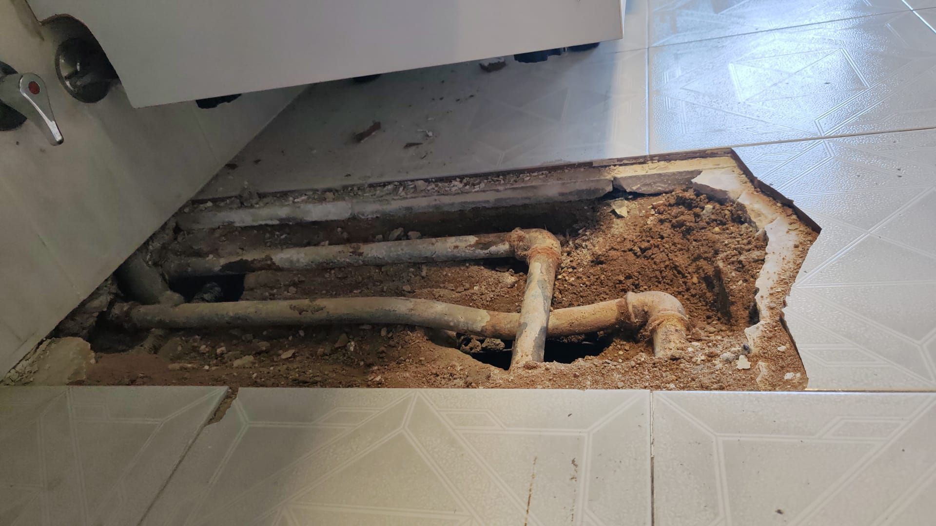 El suelo del baño está roto, dejando al descubierto tuberías y suciedad. Las baldosas están agrietadas y mojadas.