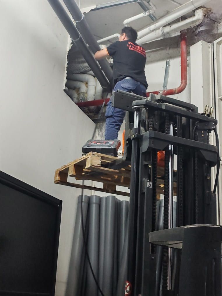 Un hombre en una plataforma de montacargas repara tuberías en un edificio. Palet de madera con tuberías negras y grises.