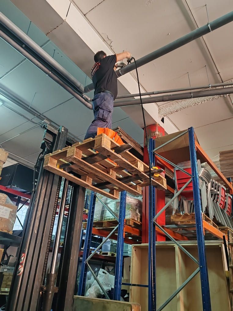 Un hombre trabaja en un palé elevado con una carretilla elevadora sobre un tubo del techo en un almacén con estanterías.
