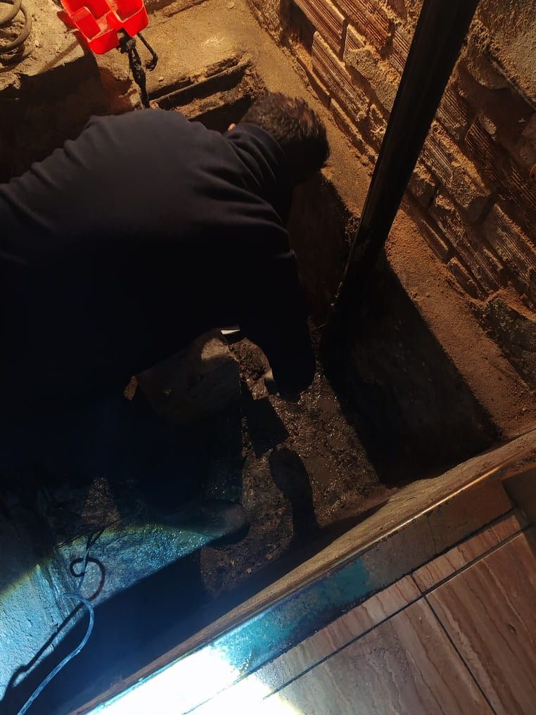 Una persona se arrodilla en un sótano oscuro, trabajando cerca de una pared de ladrillos y una tubería.