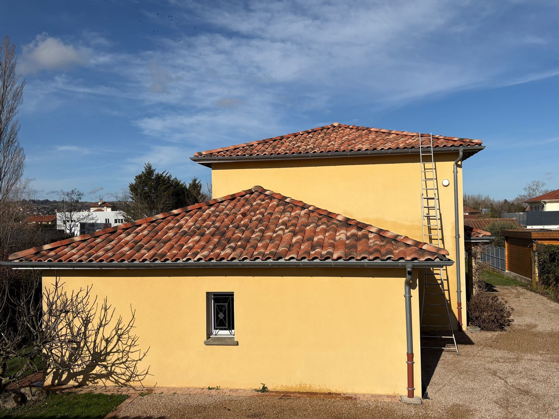 Une maison jaune à deux étages, avec un toit de tuiles en terre cuite et une échelle appuyée contre le mur extérieur.