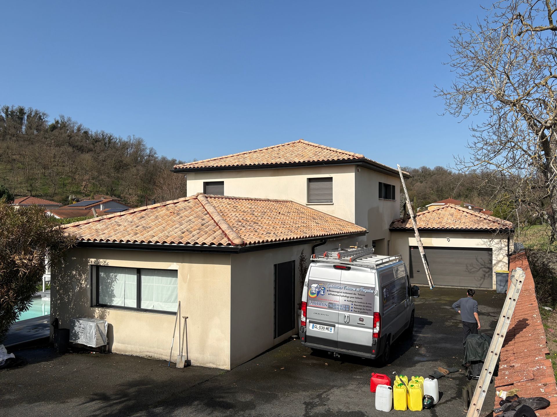 Une maison beige au toit de tuiles, avec une camionnette blanche et une personne travaillant dans l'allée.