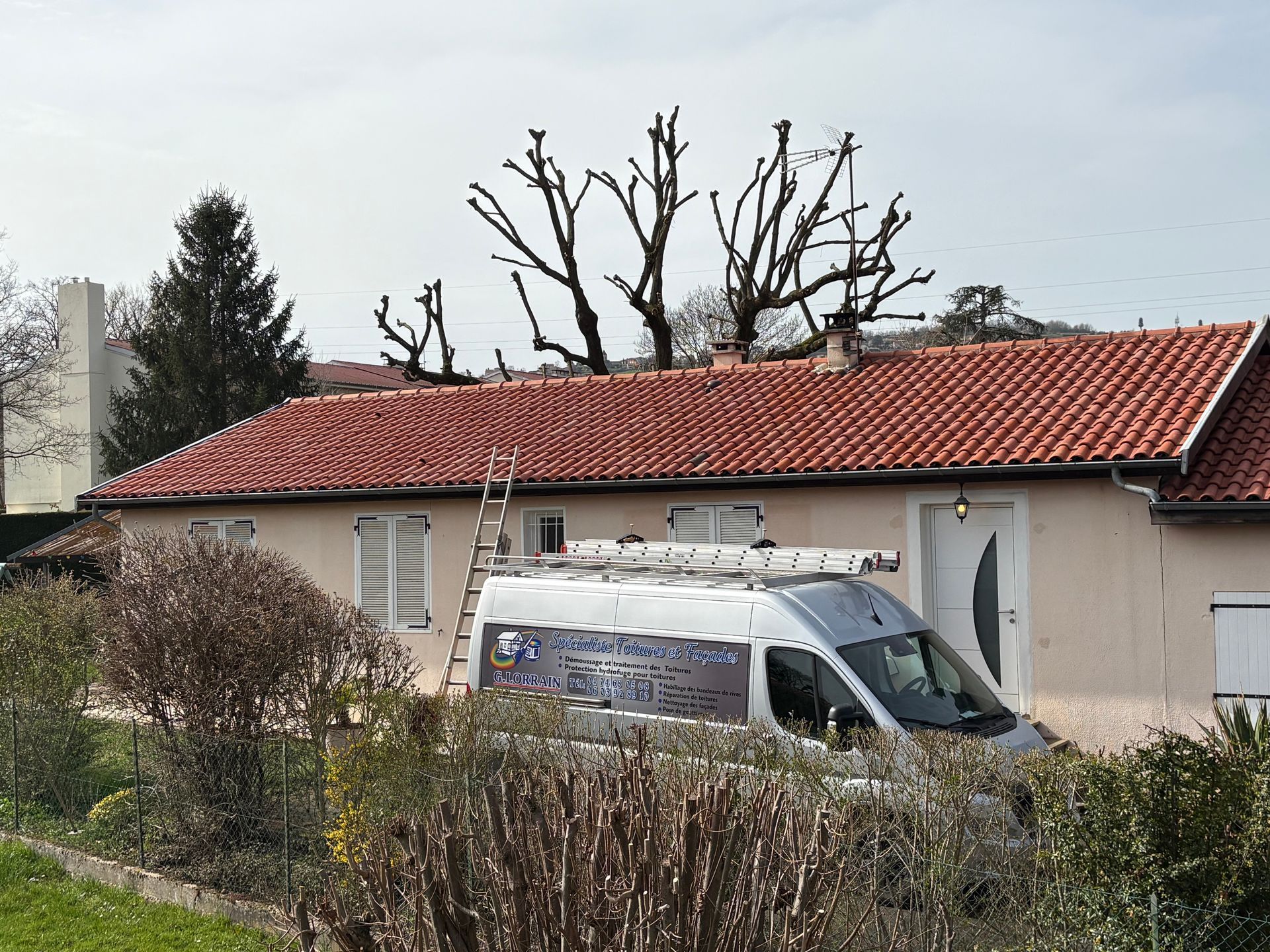 Une camionnette blanche avec une échelle sur le toit est garée devant une maison au toit de tuiles rouges et un arbre taillé.
