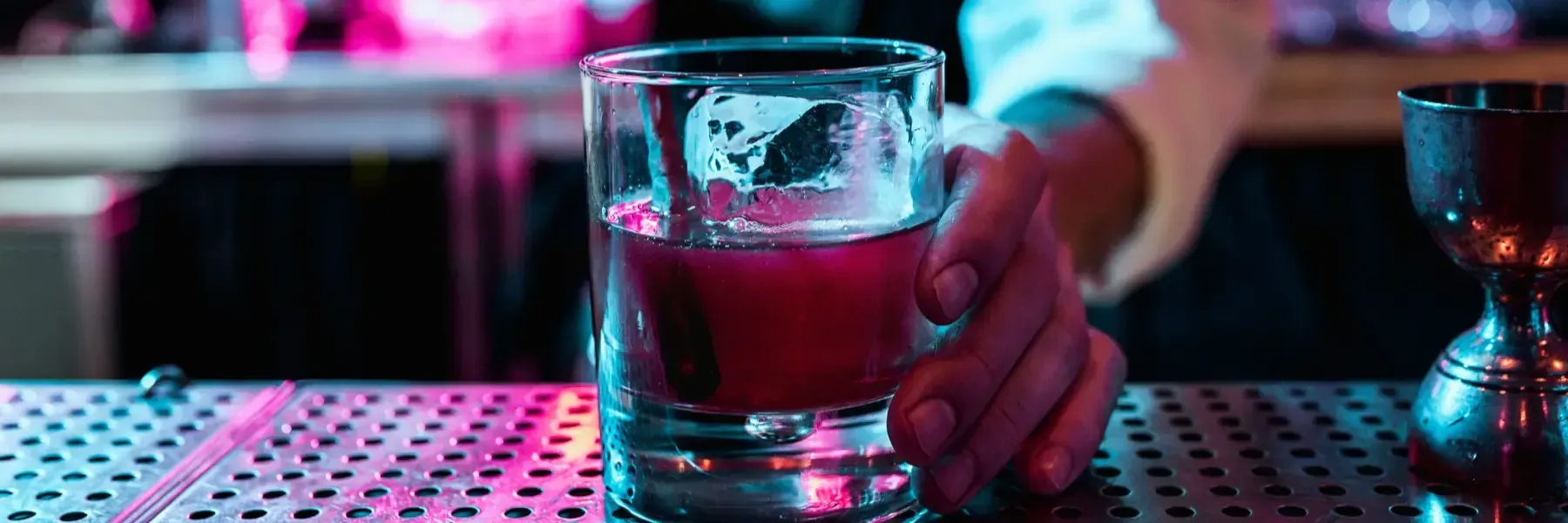 Una mano sostiene un vaso de cóctel rojo sobre la barra iluminada, con un medidor cerca, todo ello iluminado por luces
