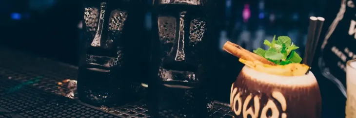 Dos tazas negras de estilo tiki se encuentran junto a un cóctel tropical servido en un coco ahuecado sobre una barra oscura.