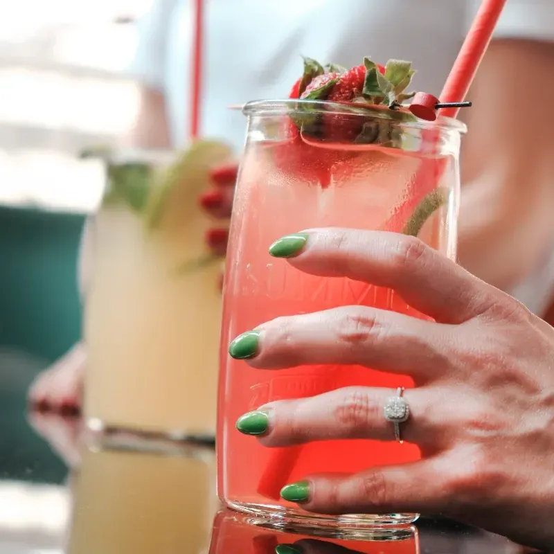 Primer plano de una mano con uñas verdes y un anillo de diamantes que sostiene un cóctel de fresa rosa en un vaso con pajita.